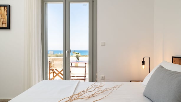 Een slaapkamer met een bed, nachtkastje, lamp, uitzicht op de zee door een schuifdeur, en een tafel met stoelen buiten.