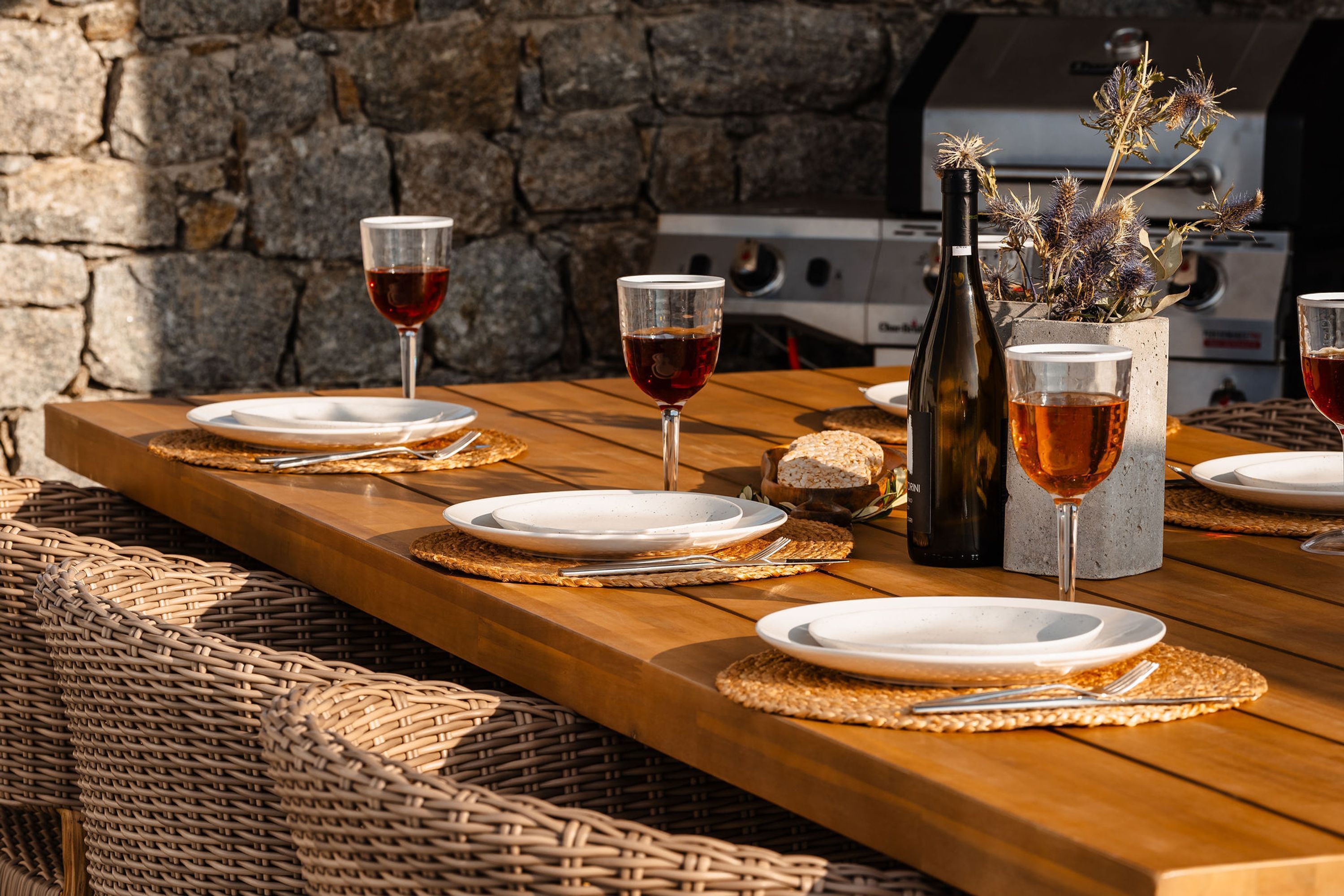 A wooden dining table set with plates, glasses, and cutlery, accompanied by wicker chairs and a stone wall in the background.