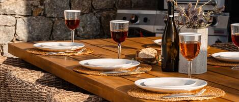 A wooden dining table set with plates, glasses, and cutlery, accompanied by wicker chairs and a stone wall in the background.