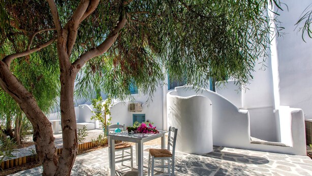 Un espace extérieur aménagé avec une table et des chaises blanches, entouré d’un sol en pierre et d’un arbre.