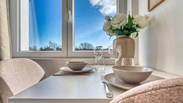 A dining table with bowls, glasses, and a vase of flowers by a window with a clear view of the sky.