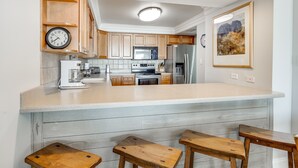 A kitchen with wooden cabinets, a large island with stools, a clock, and a framed picture on the wall. - Beach House 101D - Gulf Front Resort! (Miramar Beach)