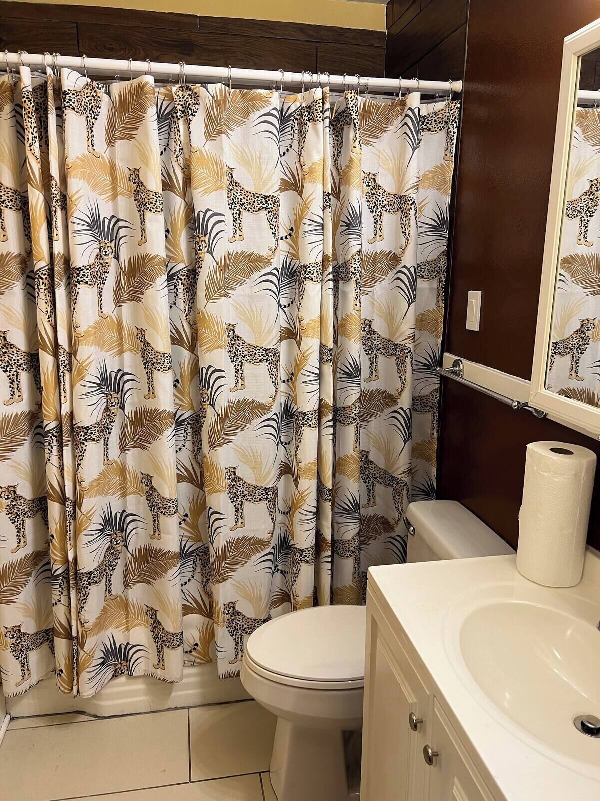 Un baño con cortina de ducha estampada con motivos de leopardo y hojas de palma, un inodoro, un lavabo y un espejo.