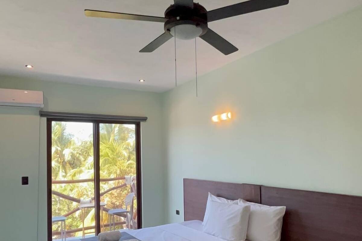 A modern hotel room with a large bed, a ceiling fan, and a view of palm trees through the sliding glass door.