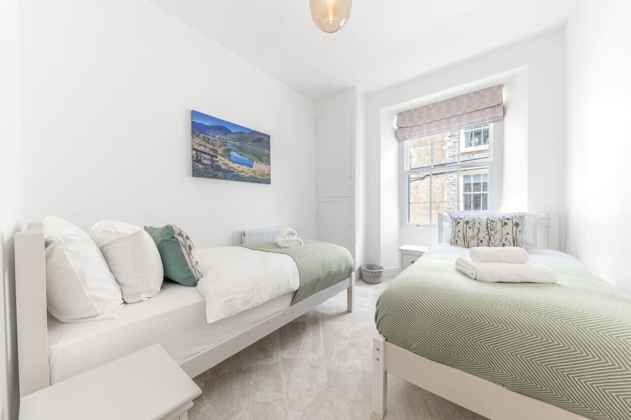 A bedroom with two beds, a window with a view of a building, and a painting on the wall.