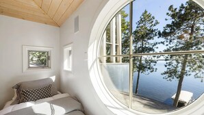 Een slaapkamer met een groot rond raam met uitzicht op een meer en bomen.