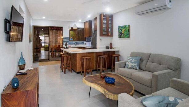 Een moderne keuken met houten kastjes, een centraal eiland en barkrukken. Er is een grijze bank en een houten salontafel met een schaal.