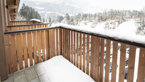 Un balcon en bois, dont la balustrade est recouverte de neige, surplombe un parking et offre une vue sur un paysage enneigé, avec des arbres et des montagnes à l’horizon.