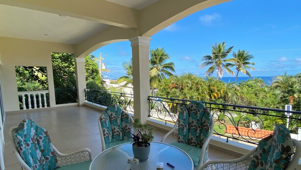 A covered outdoor seating area with a glass table, white wicker chairs with teal cushions, and a view of the ocean and palm trees. - Oceanview 5 BR w private pool, rooftop terrace & full staff 4 min from the beach (Cofresi)