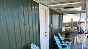 Une rangée de chaises Adirondack turquoise est placée sous un porche abrité, doté d’un toit en métal, d’une porte blanche et d’un bâtiment en métal en arrière-plan.