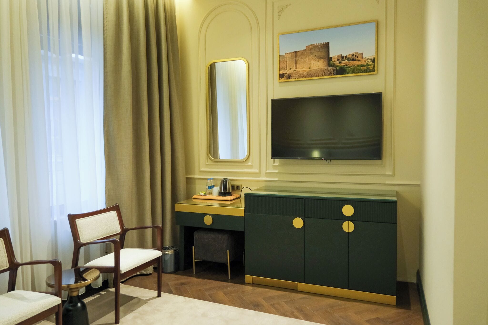 A hotel room with a wooden chair, a small table, a cabinet with a mirror, a flat-screen TV, and a painting on the wall.