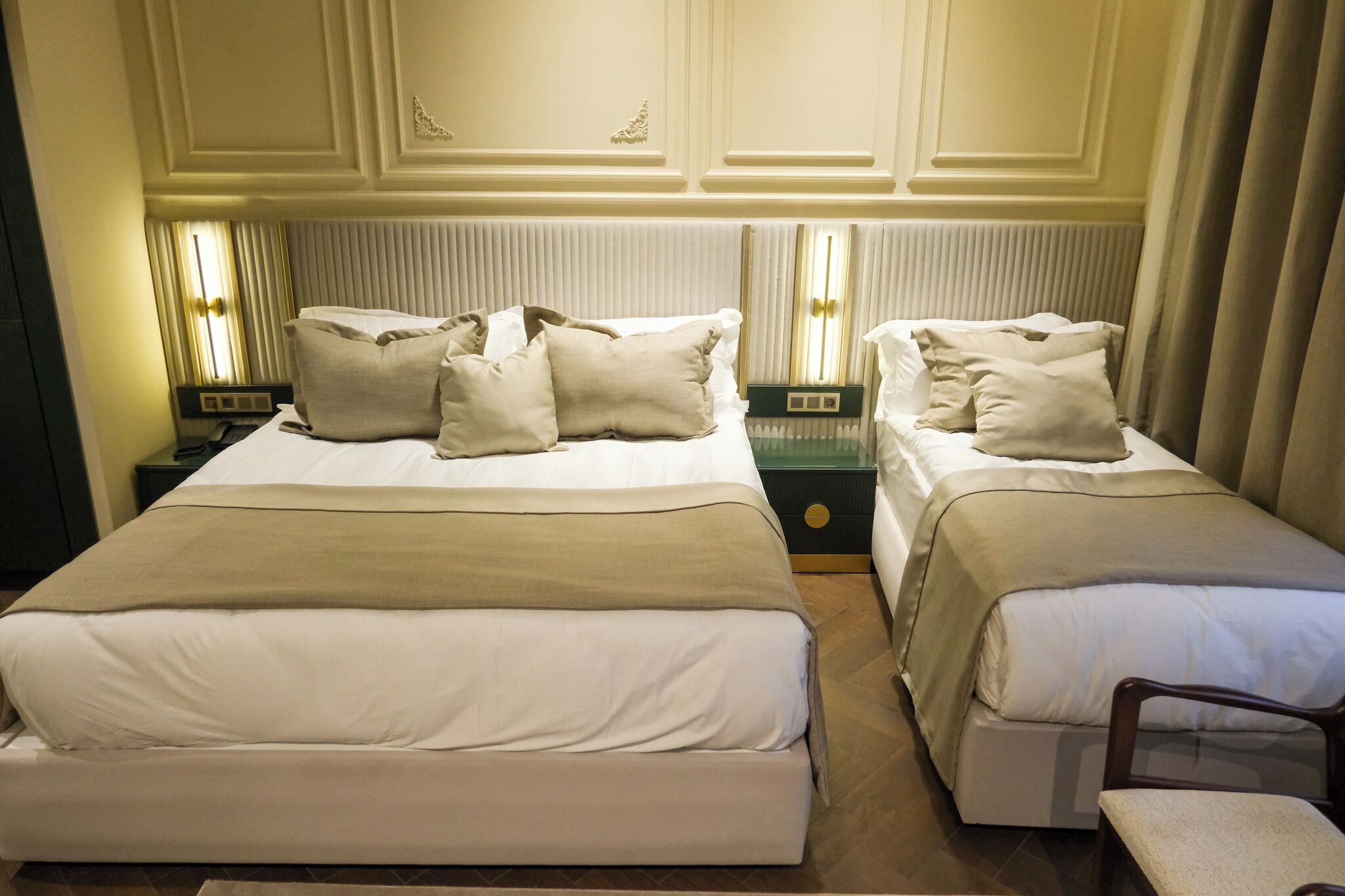 A hotel room with two beds, each with white linens and beige throw pillows. There are bedside tables with lamps, and the walls have decorative molding.