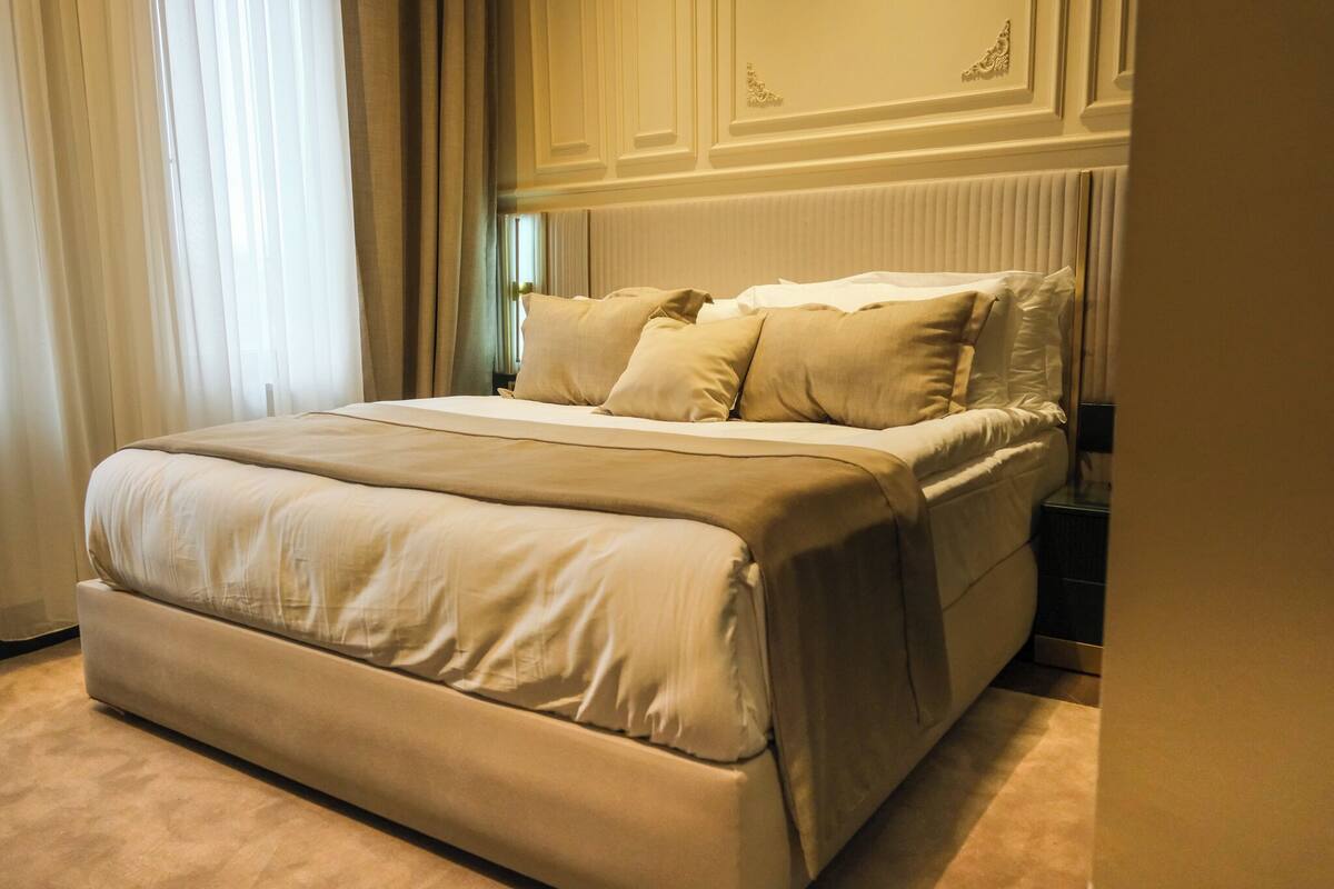 A large bed with a beige comforter and pillows, a nightstand with a lamp, and a window with sheer curtains.