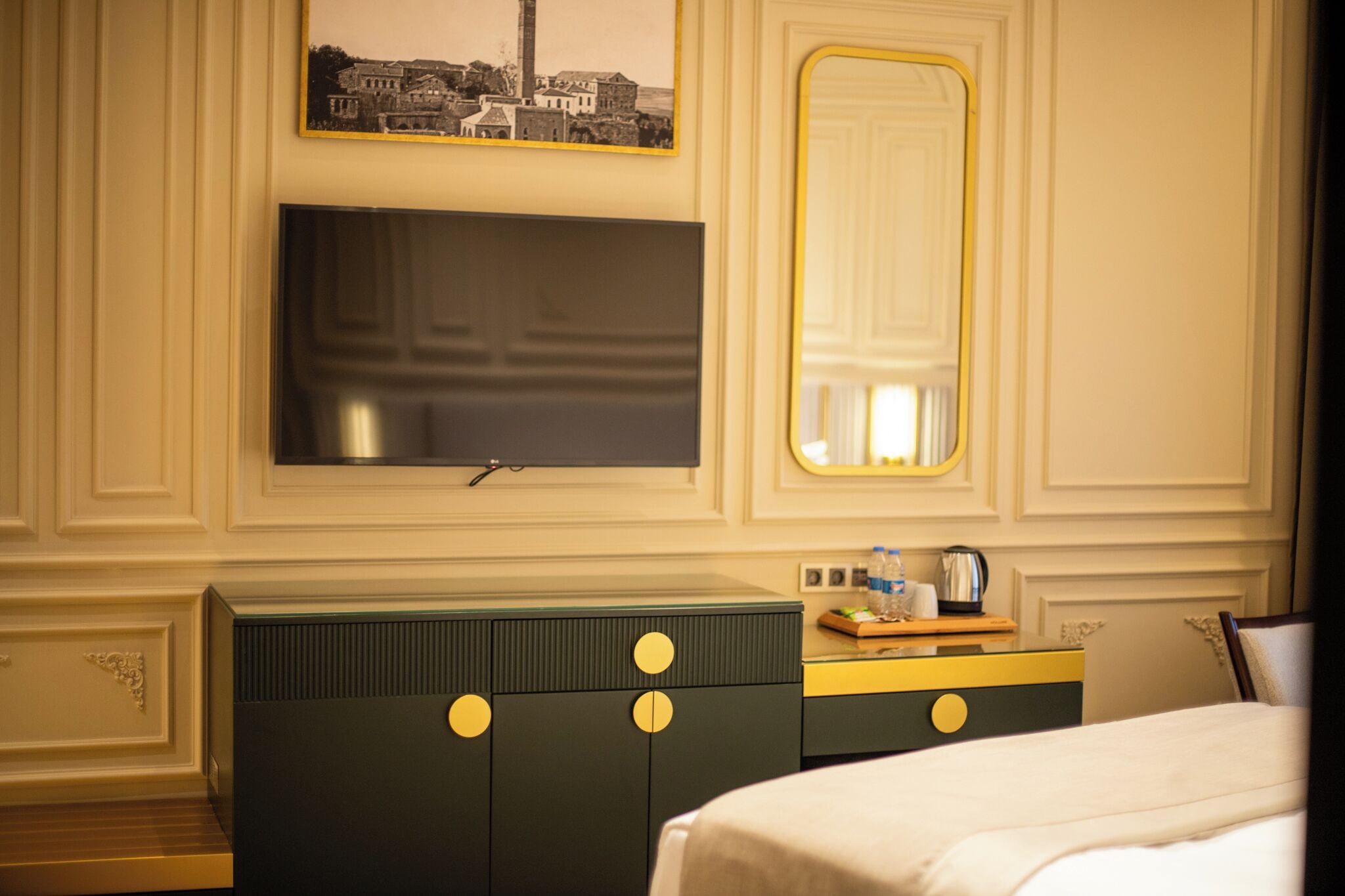 A hotel room with a large flat-screen TV mounted on the wall, a bed with white bedding, a dresser with a mirror, and a small table with a kettle and water bottles.