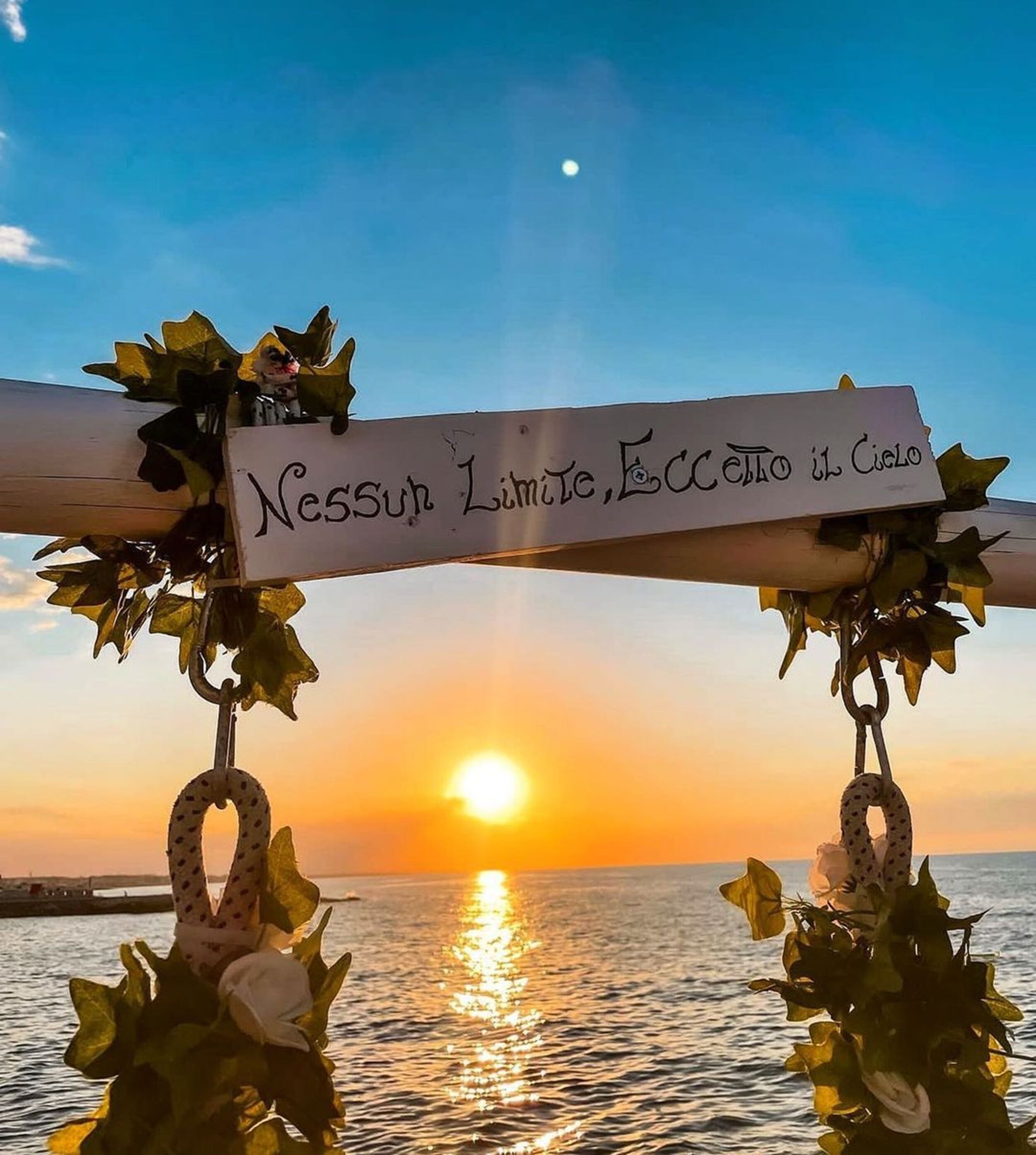 Un atardecer sobre el agua con un cartel que dice 'Nessun Limit, Eccetto il Cielo' y follaje decorativo.