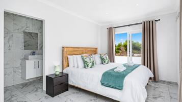 Une chambre dotée d’un grand lit, d’une table de chevet, d’une tête de lit en bois, d’une salle de bain carrelée de marbre et offrant une vue sur la verdure par la fenêtre.