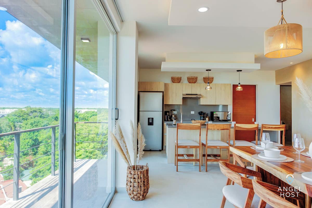 Una cocina moderna con armarios de madera, un comedor con mesa y sillas de madera, y un amplio ventanal con vistas a la vegetación.
