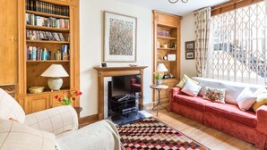 A cozy living room with a fireplace, a red sofa, a bookshelf, and a window with curtains. - Cosy Pimlico Retreat (London)