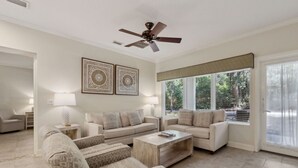 A spacious living room with a beige sofa set, a coffee table, and a ceiling fan. There are two wall-mounted lamps and a large window with blinds.
