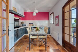 A kitchen with a glass table, blue cabinets, a pink wall, and a view through a window. - Charming 3-star Provencal villa with swimming pool and large landscaped garden (Cavalaire-sur-Mer)