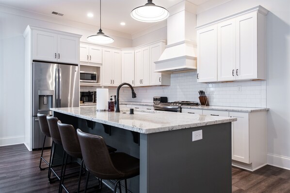 A modern kitchen with white cabinets, stainless steel appliances, and a central island with bar stools. - Watch Hill Retreat | 3BR | Norton Commons (Prospect)