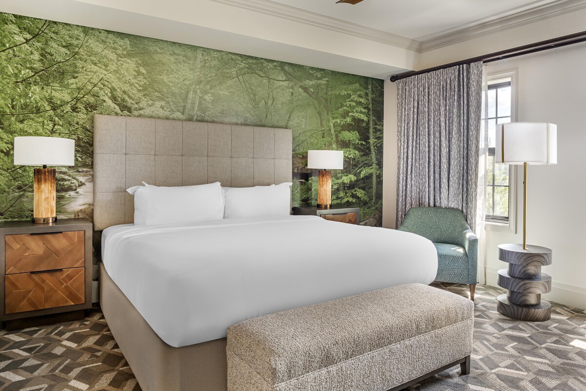 Habitación de hotel con una cama grande, cabecera, mesita de noche con lámpara, una silla y alfombra estampada.