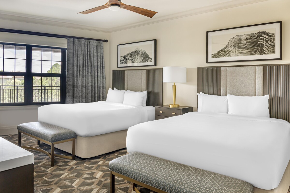 Ein Hotelzimmer mit zwei Betten, einem Schreibtisch, einer Bank, einem Deckenventilator und Bildern an den Wänden.