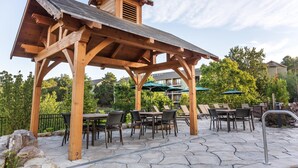 A wooden pergola with tables and chairs, surrounded by stone tiles and greenery.