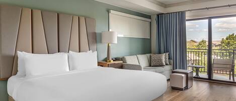 A hotel room with a large bed, a ceiling fan, a sofa, a small table, and a balcony with a view.