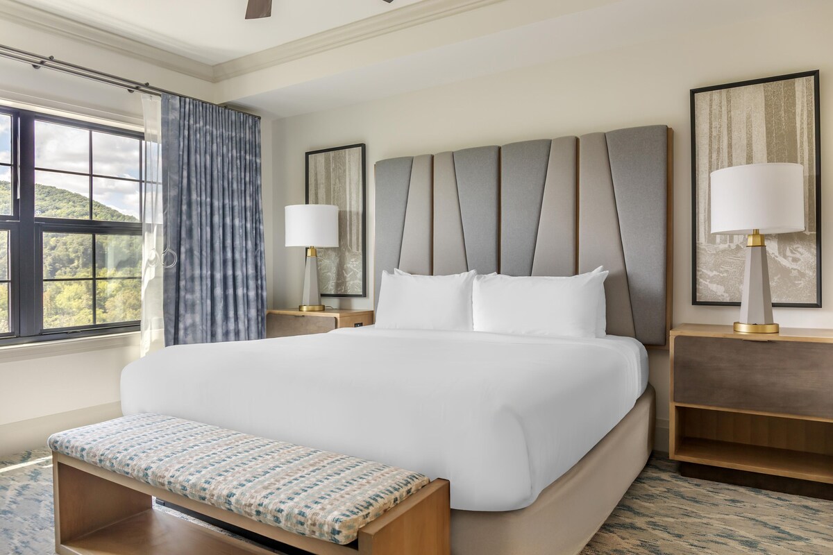 Habitación de hotel con una cama grande, mesitas de noche con lámparas, un banco y vistas al exterior.