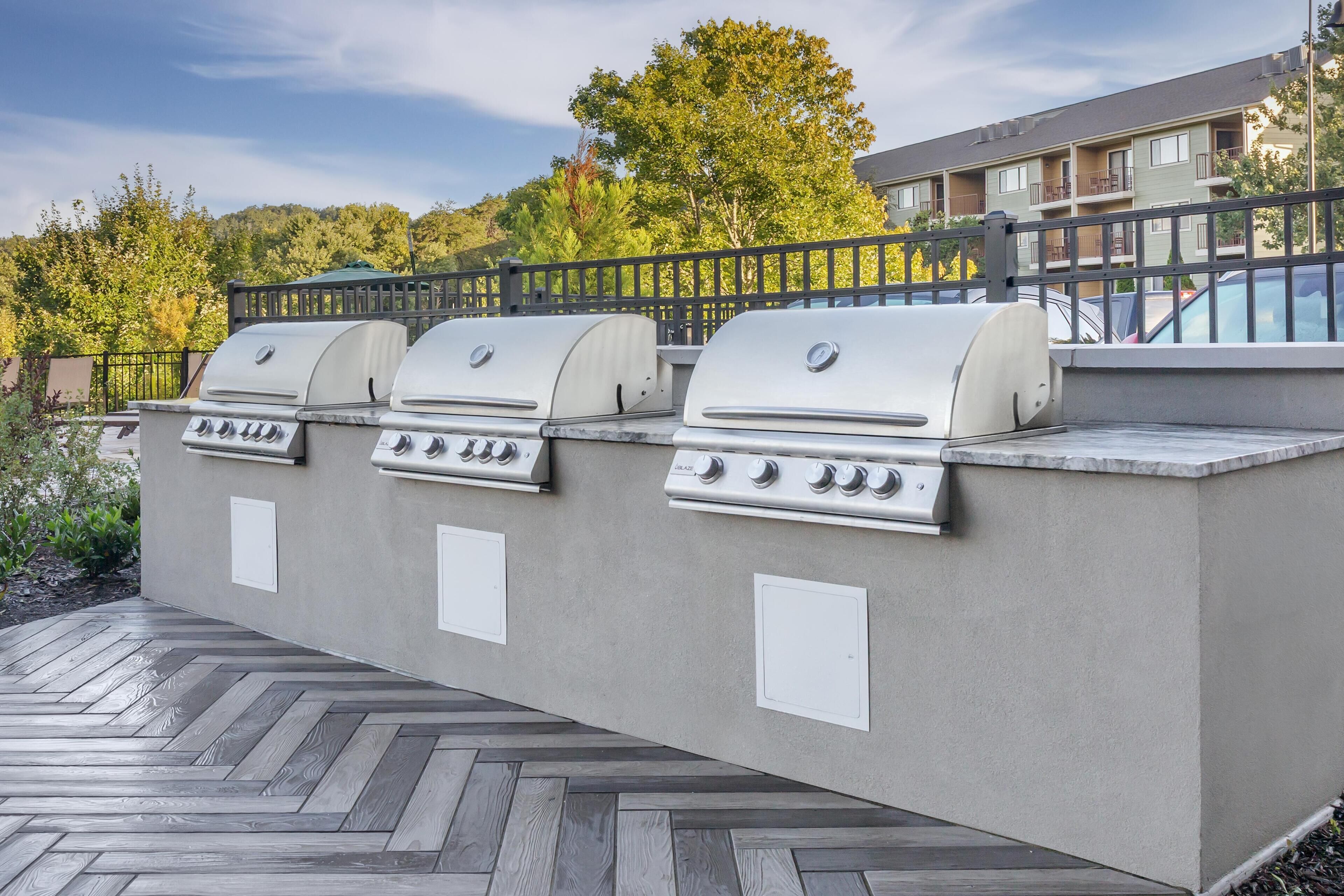 An outdoor barbecue area with three grills, a herringbone-patterned patio, and a view of a residential building in the background.