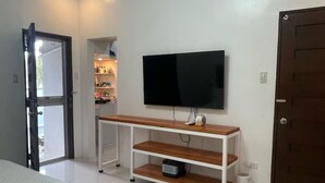 A bedroom with a bed, a TV on a wooden stand, a door, and a small trash bin. - 5BR House with Centralized AC, Backyard & Office (Roxas City)