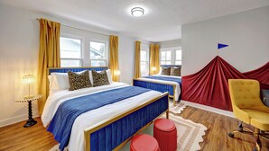 A hotel room with two beds, a red curtain, a yellow armchair, and a small table.