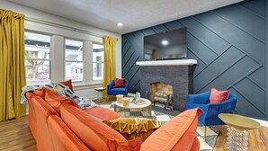 A modern living room with a sectional sofa, two blue armchairs, a fireplace, and a large TV mounted on the wall.