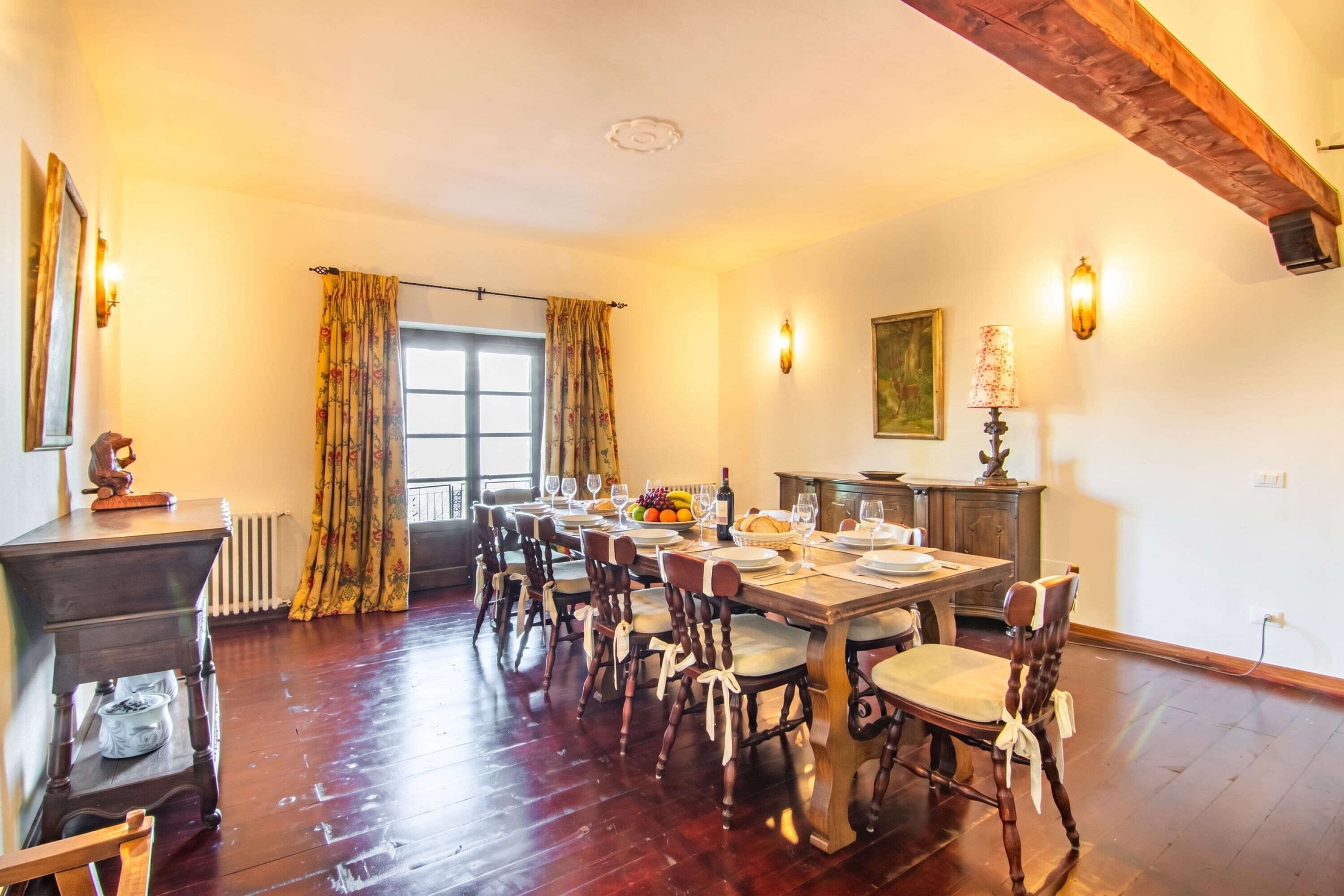 Una sala da pranzo con un tavolo in legno apparecchiato per sei persone, affiancato da sedie con sedili imbottiti. Si intravedono un termosifone e una finestra con tende.