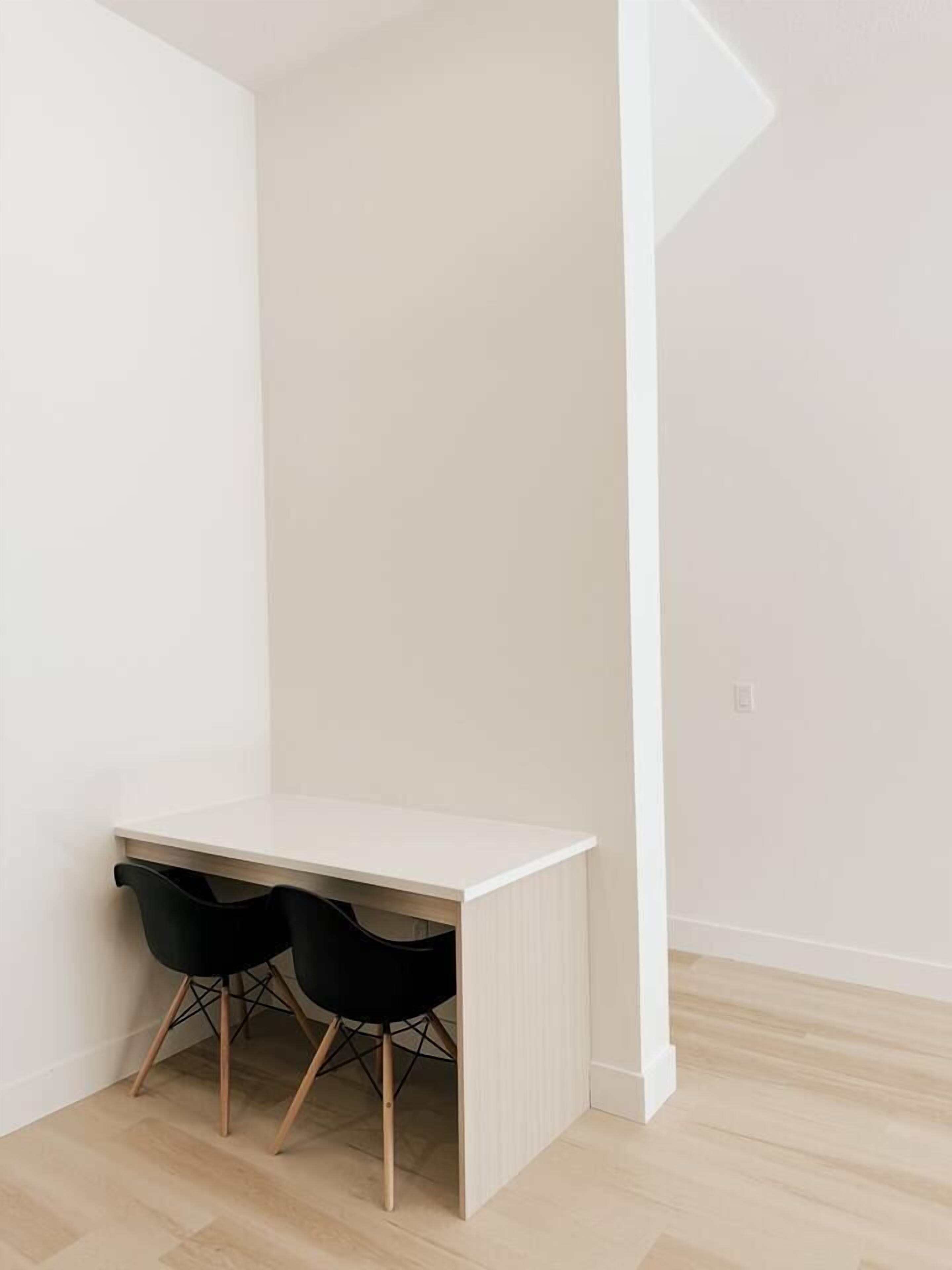 A minimalist room with a white desk and two black chairs, light wooden flooring, and white walls.