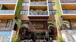 Un edificio moderno con una entrada destacada que dice "THE SHORE". Tiene fachada de madera, ventanas amplias y vegetación vertical.