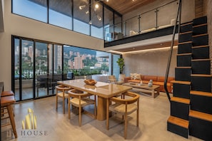 A spacious living area with a large wooden dining table, comfortable seating, a staircase, and a city view through large windows.
