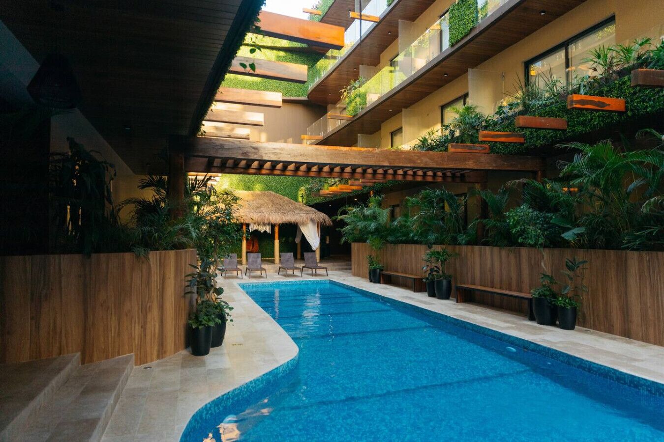 Ein Schwimmbecken mit Holzterrasse und Liegestühlen, umgeben von Topfpflanzen und einem Gazebo mit Strohdach.