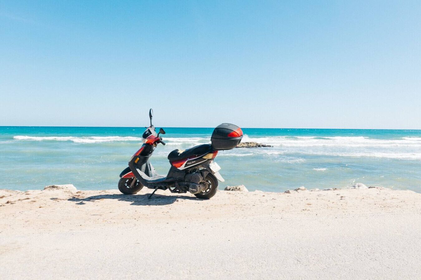 Ein Roller steht auf einem sandigen Strand, im Hintergrund das Meer.