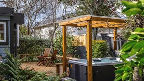 A hot tub under a wooden pergola in a backyard with a wooden deck, chairs, and lush greenery.