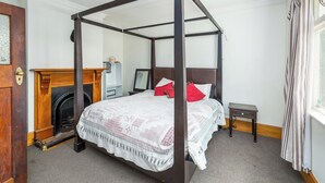A four-poster canopy bed with a wooden frame, a fireplace, a small bedside table, and a window with curtains.