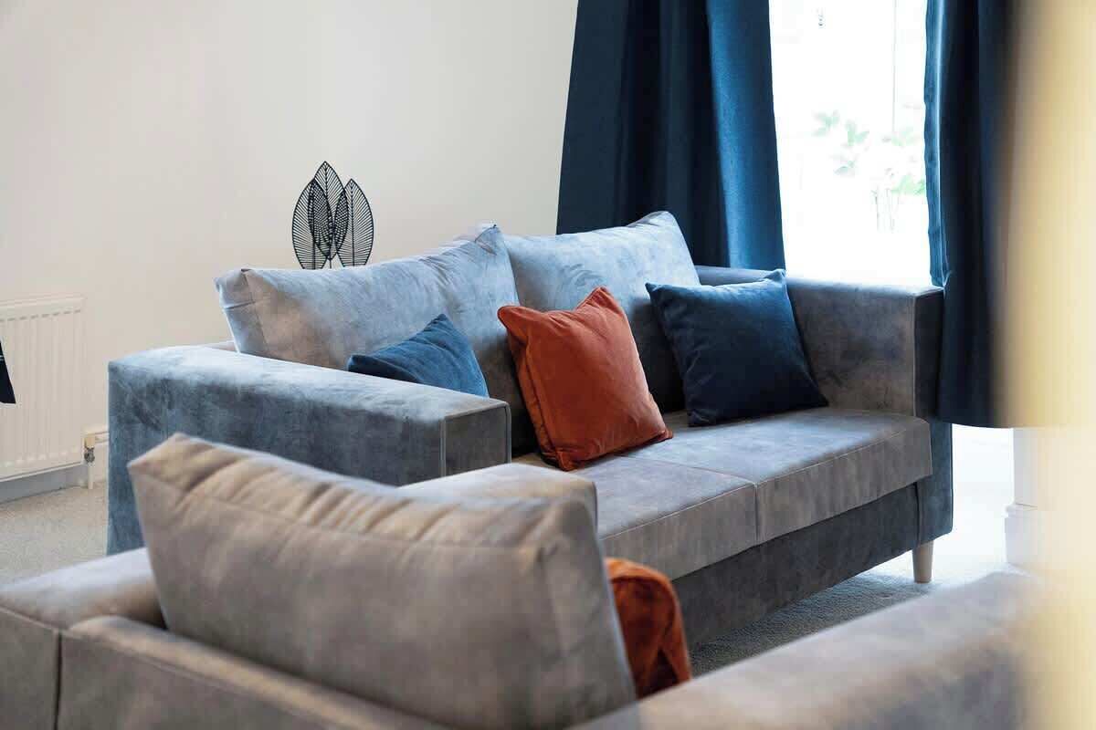 A modern living room with a grey sofa, multiple cushions, a decorative wall piece, and a window with curtains.