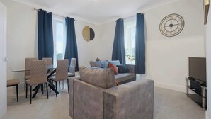 A modern living room with a sofa, dining table, chairs, a TV, and a clock on the wall.