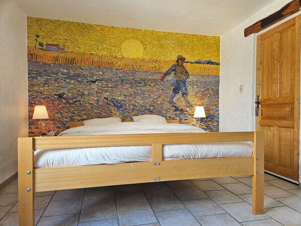 A bedroom with a bed, two bedside lamps, and a large painting of a field with a figure. - Les Grands Maitres (Montesquieu)