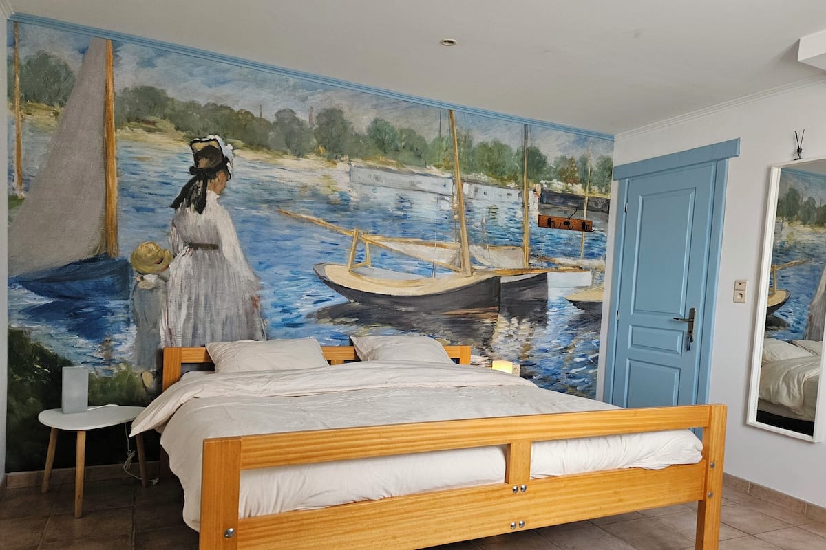 Quarto com cama de madeira, um mural com cena de rio, uma pequena mesa e uma porta azul.