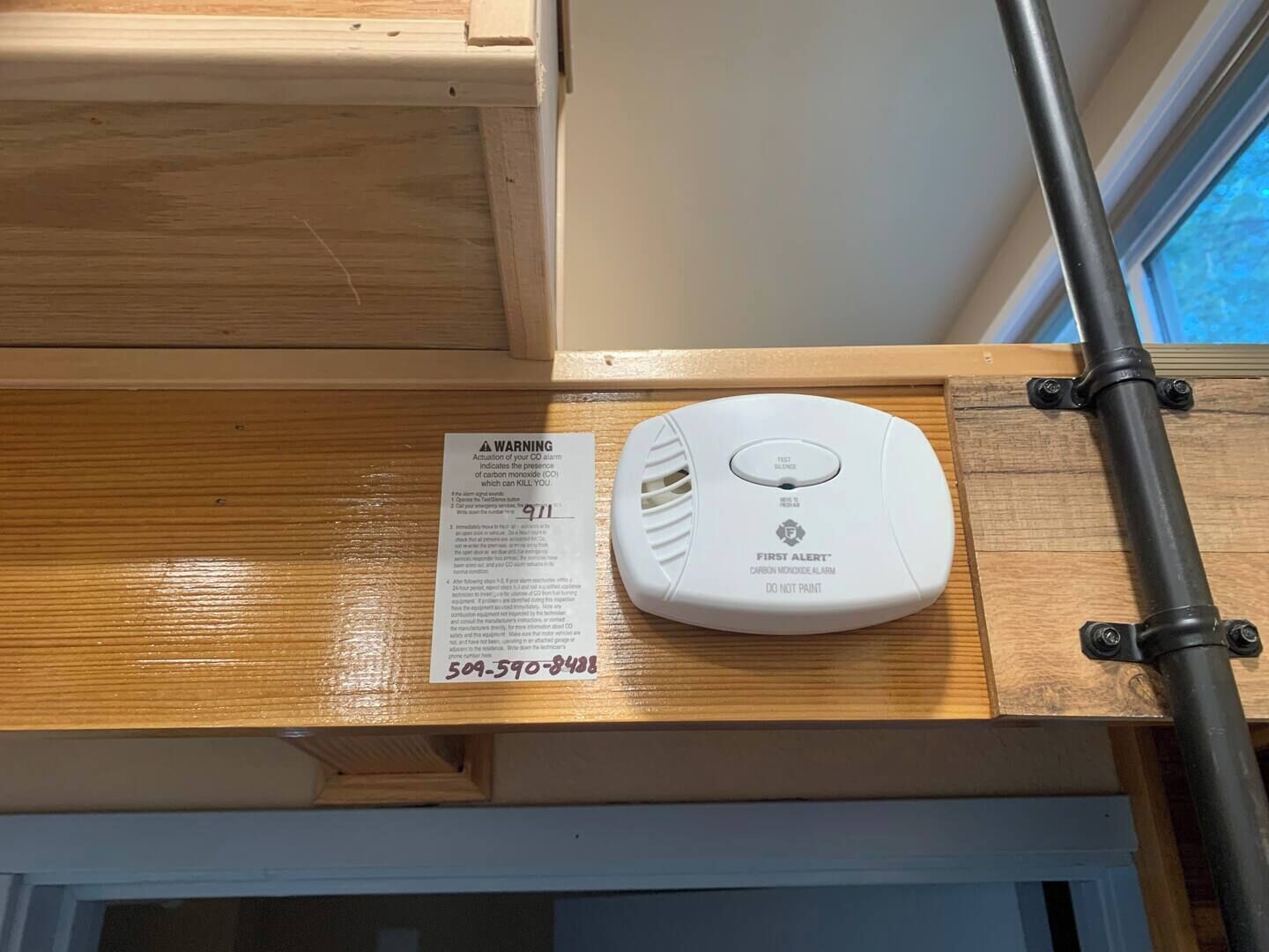 A carbon monoxide alarm mounted on a wooden shelf, with a warning label and a phone number.