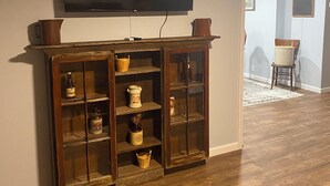 A living room with a flat-screen TV mounted on the wall, wooden shelving with various decorative items, and a wooden floor.