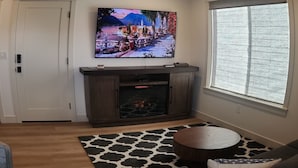 Una sala de estar con chimenea, un televisor grande montado encima y una ventana con persianas, además de una alfombra estampada.
