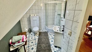 Un baño con inodoro, bidé y ducha. El suelo tiene un diseño de cuadros, y hay una toalla sobre una silla.